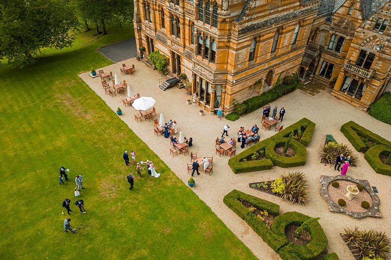 Aerial view of a wedding reception on the grounds of a large, historic building. Guests are gathered on a patio and lawn, some seated at tables and others standing. The garden features neatly trimmed hedges and manicured lawns. Image by Documentary Wedding Photographer Lewis Bishop Photography.
