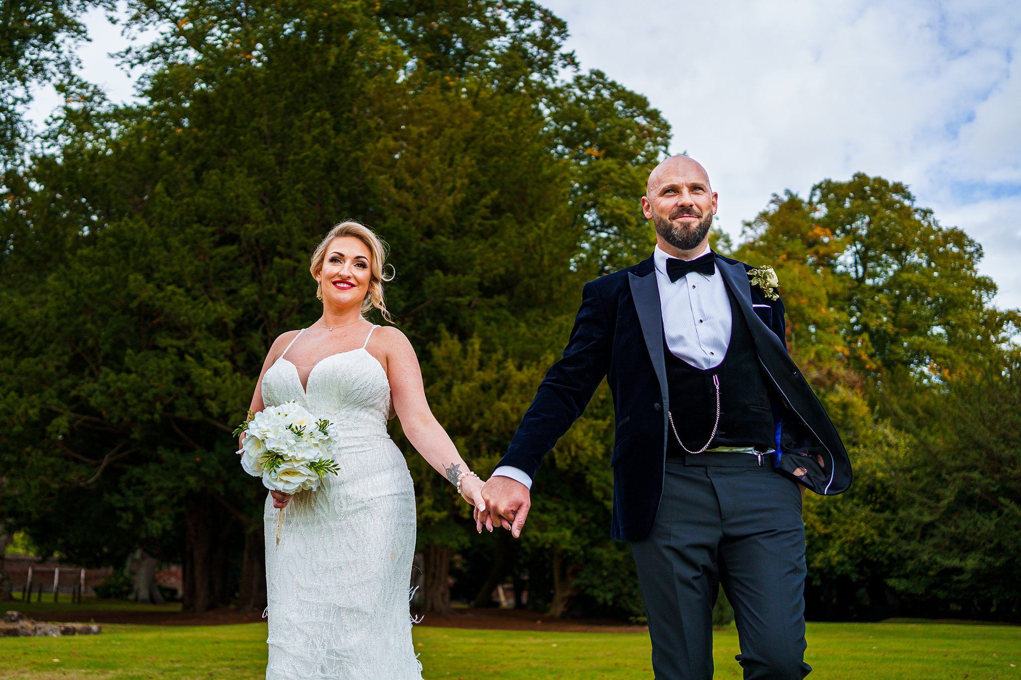 A bride in a white wedding dress and a groom in a black tuxedo hold hands while walking on a lush green lawn. The bride is holding a bouquet of white flowers, and they are both smiling. Tall trees and a cloudy sky are visible in the background. Image by Documentary Wedding Photographer Lewis Bishop Photography.