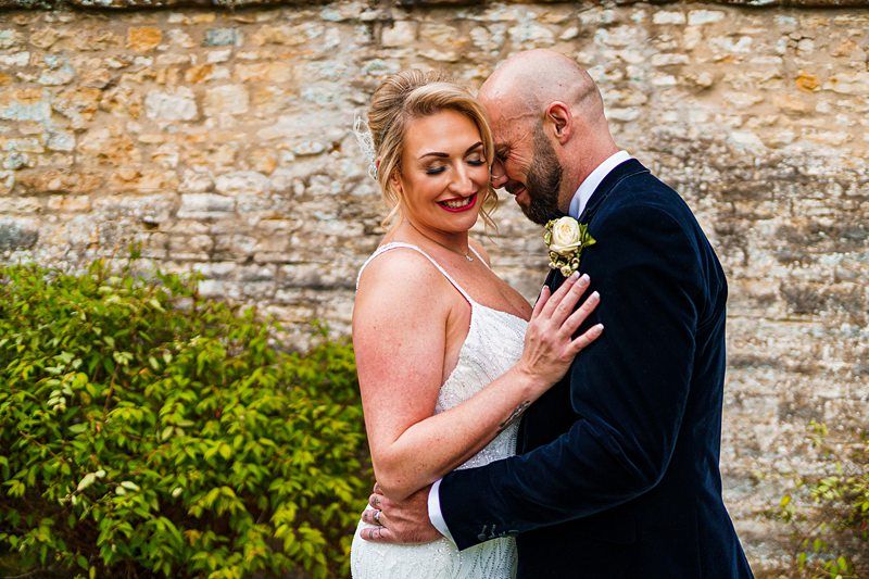 A bride and groom stand close together in front of a stone wall, both smiling contentedly. The bride, in a white lace dress, rests her hand on the groom's chest. The groom, in a dark suit with a white rose boutonniere, leans his forehead against hers. Image by Documentary Wedding Photographer Lewis Bishop Photography.
