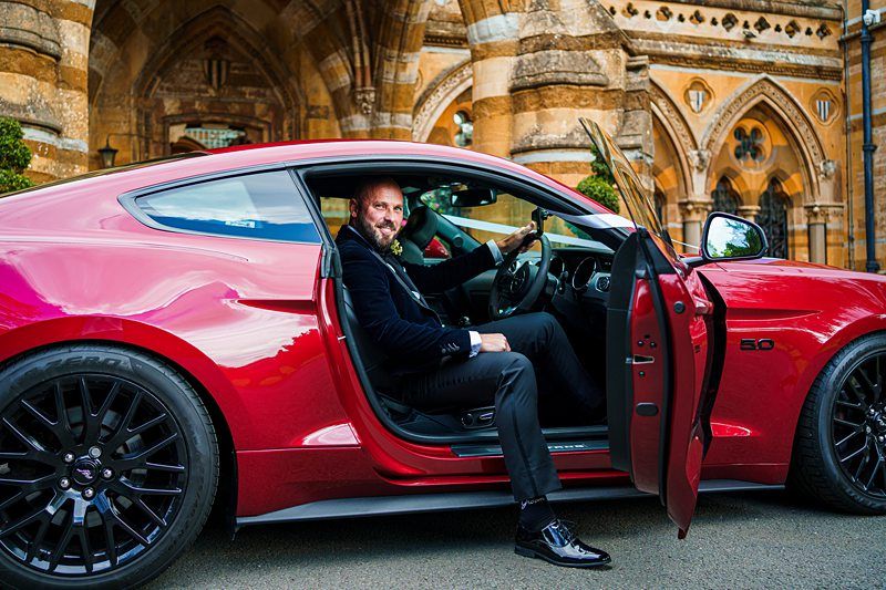A man sitting in a red car. Image by Documentary Wedding Photographer Lewis Bishop Photography.