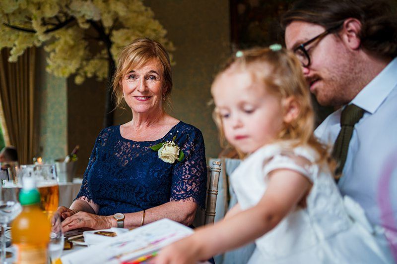 A woman in a blue dress with a white flower corsage sits at a table, smiling at the camera. Next to her, a man with glasses and a child in a white dress are engaged in an activity. The table holds drinks and papers, and a lush decoration is in the background. Image by Documentary Wedding Photographer Lewis Bishop Photography.