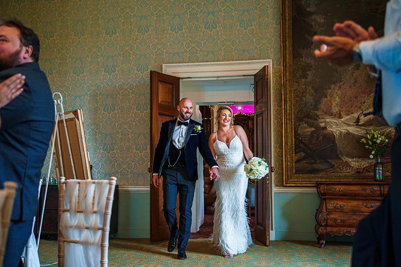 A joyful couple enters a beautifully decorated room hand in hand, smiling. The groom is bald with a beard, wearing a dark suit, while the bride wears a white dress holding a bouquet. Guests in the foreground appear to applaud, and a large painting adorns the wall. Image by Documentary Wedding Photographer Lewis Bishop Photography.