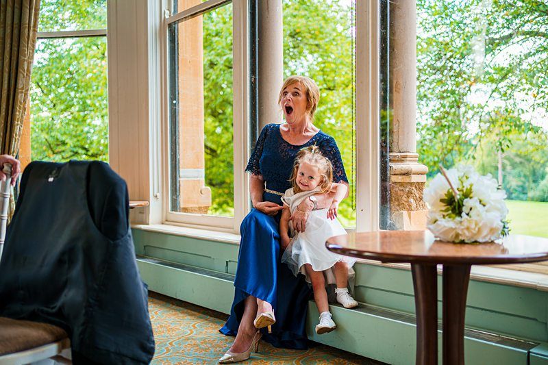 A woman in a blue dress, appearing surprised with her mouth open, sits on a ledge by large windows, holding a smiling girl in a white dress. They are next to a small round table with a bouquet of white flowers. Green trees are visible outside the windows. Image by Documentary Wedding Photographer Lewis Bishop Photography.