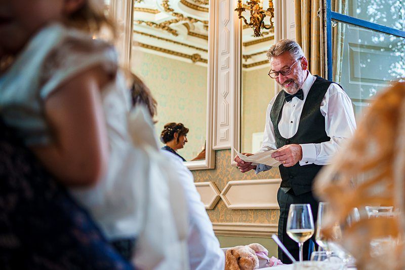 An older man in a black vest and bow tie stands and reads from a piece of paper, likely giving a speech. The setting is a formal, ornate room with mirrors and chandeliers. Blurred in the foreground are seated guests and a child. Wine glasses and table decor are visible. Image by Documentary Wedding Photographer Lewis Bishop Photography.