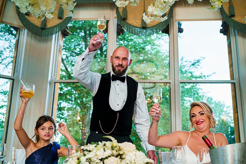 A groom in a black vest and bow tie toasts with a champagne glass, accompanied by a bride in a white gown and a young girl in a blue dress, all raising their glasses in celebration. They are indoors, with large windows and floral decor in the background. Image by Documentary Wedding Photographer Lewis Bishop Photography.