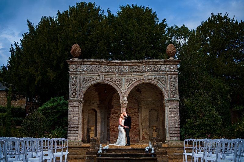 A couple dressed in wedding attire stands under a stone archway adorned with intricate carvings. They are embraced, surrounded by lush greenery and rows of white chairs arranged for a ceremony. The sky above is partly cloudy, adding to the romantic atmosphere. Image by Documentary Wedding Photographer Lewis Bishop Photography.