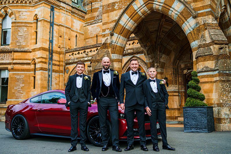 Four men dressed in black tuxedos stand in front of a red sports car and a large, ornate stone building. The group appears to be in formal attire for a special occasion. Two of the men have flower boutonnieres on their lapels. Image by Documentary Wedding Photographer Lewis Bishop Photography.