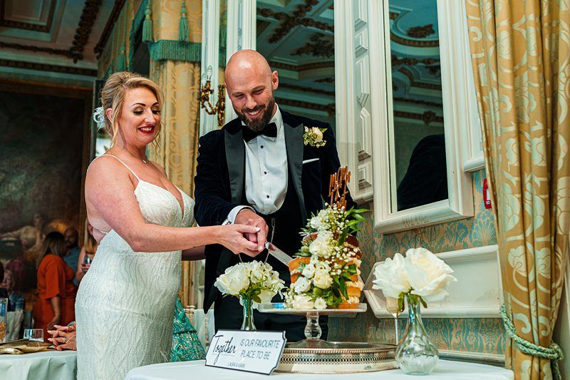 A joyous bride and groom in elegant attire are cutting a white wedding cake adorned with flowers. The setting appears to be an opulent, ornately decorated room with large mirrors and intricate details. White flowers are arranged in vases on the table. Image by Documentary Wedding Photographer Lewis Bishop Photography.