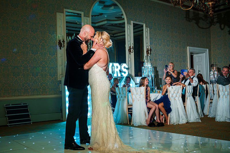 A bride and groom embrace during their first dance at a wedding reception. They are on a white dance floor with chairs and tables in the background. Guests sit and watch, some smiling. A sign with illuminated letters "MR & MRS" is visible behind the couple. Image by Documentary Wedding Photographer Lewis Bishop Photography.