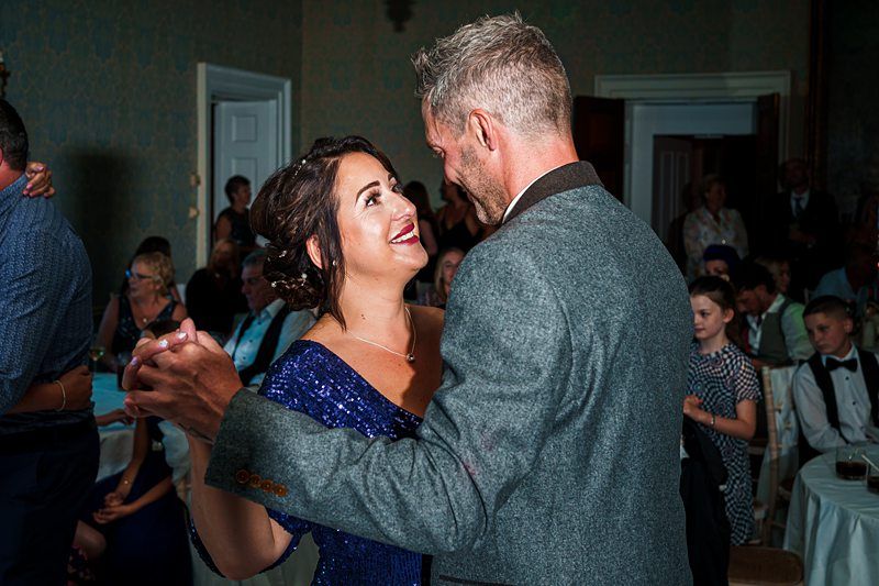 A couple dressed in formal attire shares a dance at a wedding reception. The woman is wearing a sparkly blue dress while the man is in a gray suit. They are smiling at each other with guests seated and standing in the background. Image by Documentary Wedding Photographer Lewis Bishop Photography.