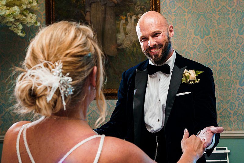 A bald man with a beard, wearing a dark tuxedo and a bow tie, smiles warmly at a woman with blonde hair styled in an updo with a white hair accessory. The woman, in a white dress, has her back to the camera. They are indoors with elegant wallpaper in the background. Image by Documentary Wedding Photographer Lewis Bishop Photography.