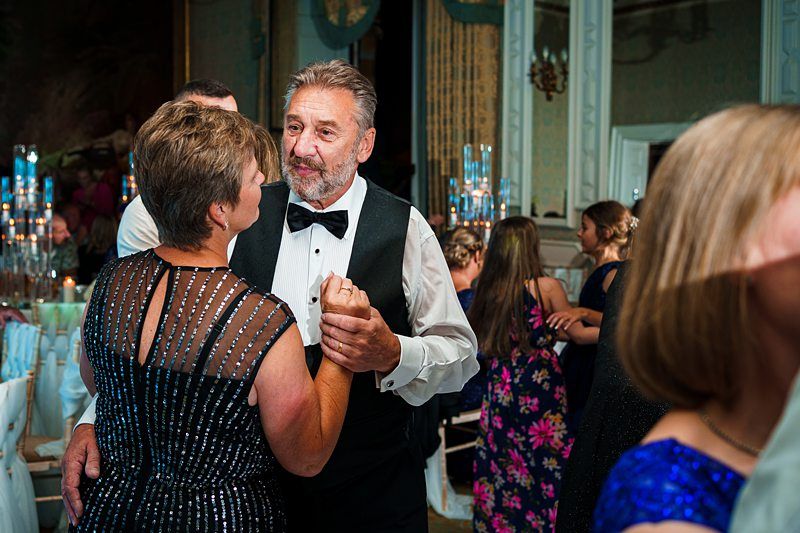 A man in a black tuxedo and bow tie is dancing with a woman in a sparkly dress at a formal event. Other attendees can be seen dancing and mingling in the background. The setting appears to be an elegant, well-lit ballroom adorned with candles and decorations. Image by Documentary Wedding Photographer Lewis Bishop Photography.