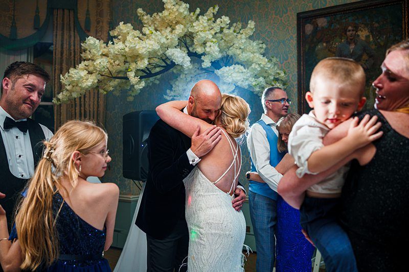 A bride and groom share a hug surrounded by family and friends at a wedding reception. They are in an elegant room with floral decorations. A young boy is being held by a woman on the right, and several guests in formal attire are visible in the background. Image by Documentary Wedding Photographer Lewis Bishop Photography.
