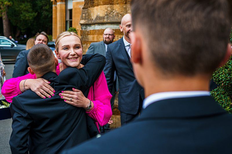 A woman in a bright pink outfit warmly embraces a young boy outside a building, as several people in suits look on and smile. Another young boy in a suit stands in the foreground, facing the embracing pair. A parked car and greenery are visible in the background. Image by Documentary Wedding Photographer Lewis Bishop Photography.