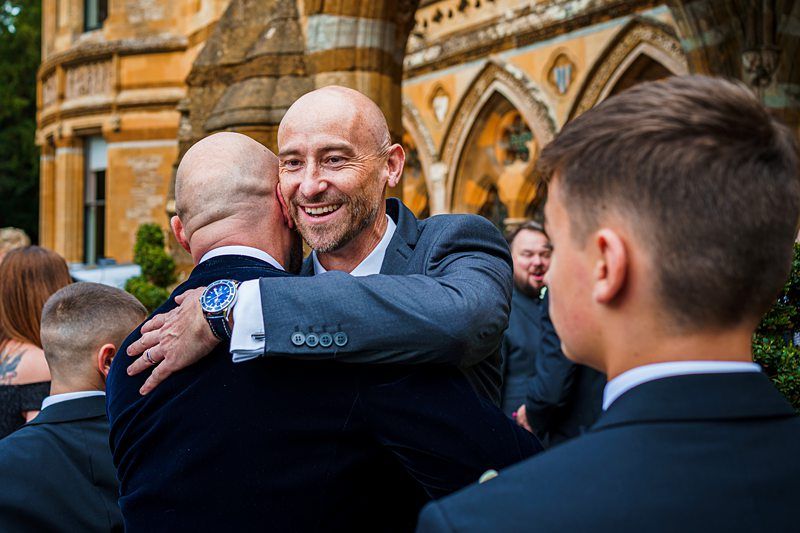 A bald man in a suit warmly hugs another man at an outdoor event, with a historic building in the background. Other people, including a boy, are nearby, dressed formally. The atmosphere appears joyful and celebratory. Image by Documentary Wedding Photographer Lewis Bishop Photography.