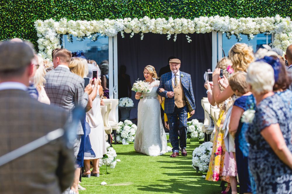 A smiling bride in a white dress and veil is walking down the aisle with an older man in a checkered suit and hat. They are outdoors, surrounded by guests clapping and taking photos, with a backdrop of green foliage and white floral arrangements. Image by Documentary Wedding Photographer Lewis Bishop Photography.