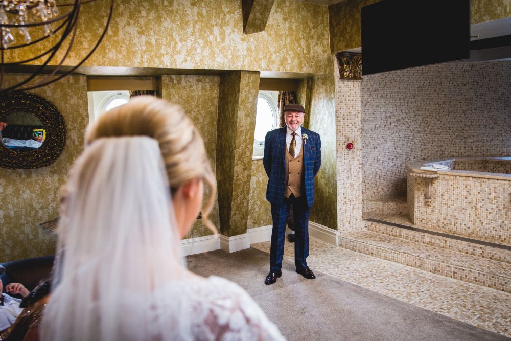 A bride in a white dress and veil is turned towards an older man in a blue checkered suit and brown tie, who is standing and holding a cane inside a warmly lit room. The room features textured golden walls, circular windows, and a mosaic-tiled area with steps. Image by Documentary Wedding Photographer Lewis Bishop Photography.