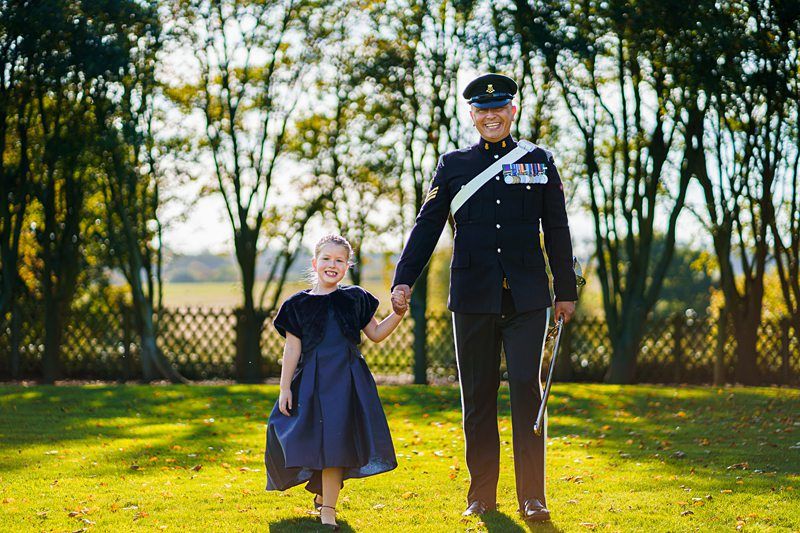A man in a formal military uniform smiles while holding hands with a young girl in a dark blue dress. They are standing on grass, with tall trees and a sunny sky in the background. The man has a sword by his side, and they both appear happy. Image by Documentary Wedding Photographer Lewis Bishop Photography.