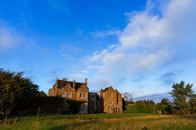 A sprawling stone mansion with numerous gables and chimneys stands in a grassy field under a bright blue sky with scattered clouds. The house is partially covered in ivy, and there are trees and bushes surrounding the property. Image by Documentary Wedding Photographer Lewis Bishop Photography.