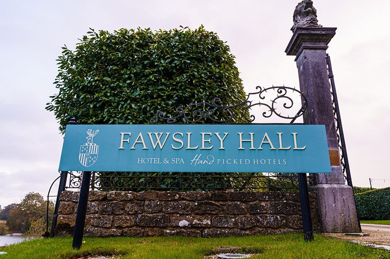 A sign for Fawsley Hall Hotel & Spa, Hand Picked Hotels, stands in front of a trimmed hedge. The sign is teal with white and gold lettering and a crest. The base is made of stone, and a decorative black metal frame surrounds the sign, with a stone pillar on the right. Image by Documentary Wedding Photographer Lewis Bishop Photography.