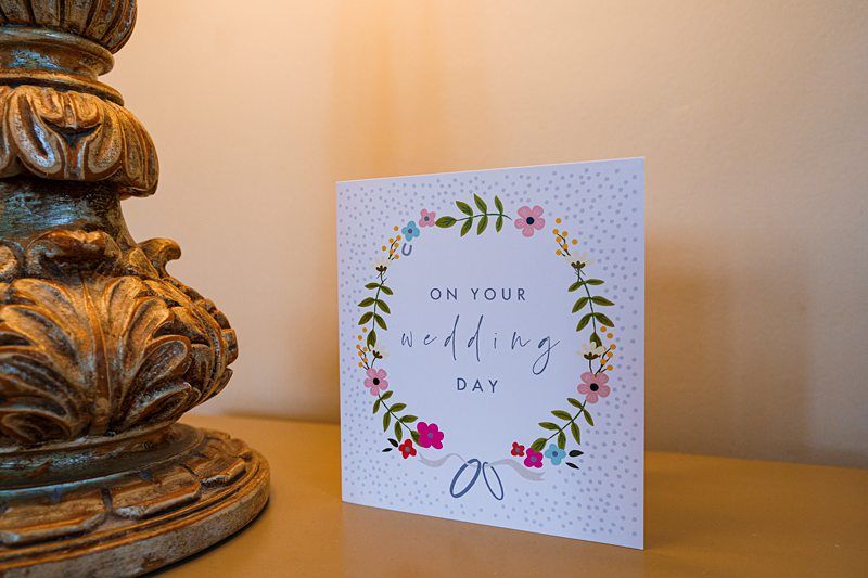 A wedding card with a floral wreath design and the words "On Your Wedding Day" is displayed upright on a shelf next to an ornate, decorative lamp base. The card features flowers and leaves in various colors and a small blue bow at the bottom of the wreath. Image by Documentary Wedding Photographer Lewis Bishop Photography.