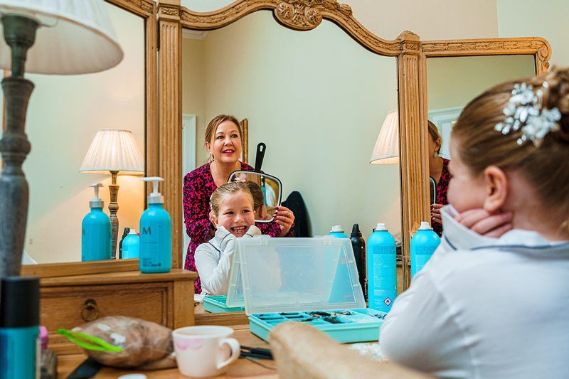 A young girl smiles while sitting at a vanity table with her hair styled for a special occasion. Behind her, an adult holds a curling iron. The reflection shows hair products and jewelry boxes on the table. The room is warmly lit with beige walls and a wooden-framed mirror. Image by Documentary Wedding Photographer Lewis Bishop Photography.