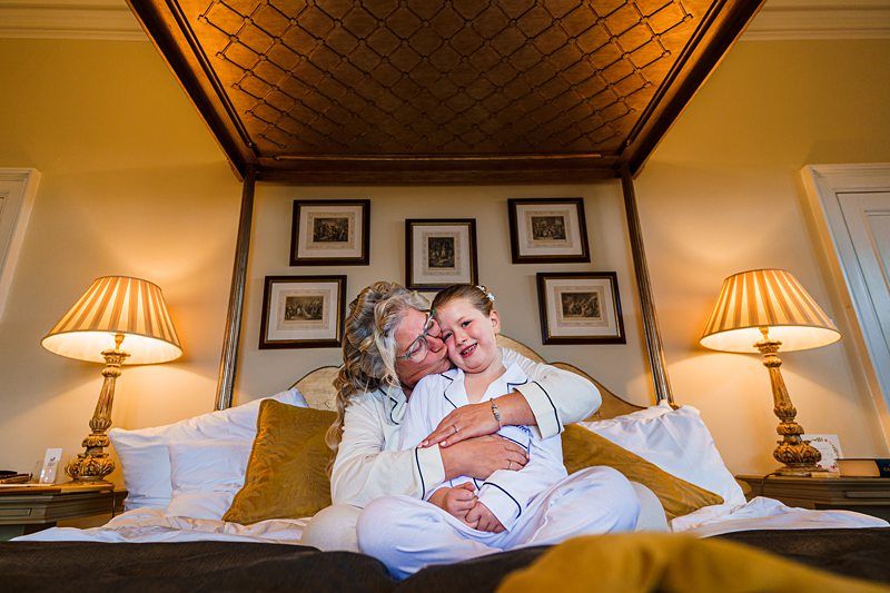 A person with long hair embraces a smiling child on a bed under a canopy. The room features warm lighting from two bedside lamps and framed pictures on the wall. Both individuals are dressed in light-colored pajamas. Image by Documentary Wedding Photographer Lewis Bishop Photography.
