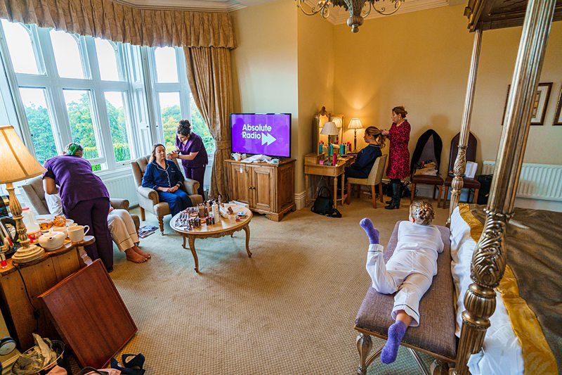 A group of people in a cozy, well-lit room with large windows. Some are engaged in conversations while others sit or lie down. A TV is on displaying the Absolute Radio logo. There are beauty products on a table, indicating a relaxed, social gathering. Image by Documentary Wedding Photographer Lewis Bishop Photography.