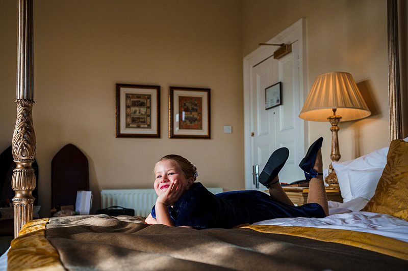 A young child wearing a blue dress lies on their stomach on a bed in a warmly lit room, resting their chin on their hands with a smile. The room features a tall lamp, framed pictures on the wall, and ornate bedposts. The child looks relaxed and happy. Image by Documentary Wedding Photographer Lewis Bishop Photography.