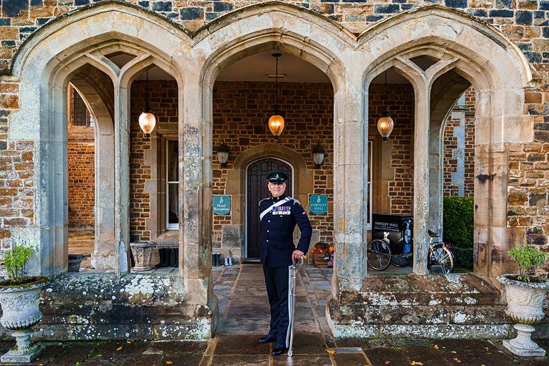 A uniformed guard stands at attention under a stone archway of a historic building. The structure features three arches, hanging lamps, potted plants, and a bicycle parked to the side. The building's stone facade and partially visible interior add to the historic ambiance. Image by Documentary Wedding Photographer Lewis Bishop Photography.