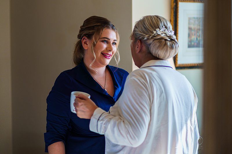 A woman with a blue dress and rolled-up sleeves is holding a white mug and smiling at another woman with blonde hair in an updo, wearing a white shirt with a pearl hair accessory. They are standing indoors in a warmly lit room with a framed picture on the wall. Image by Documentary Wedding Photographer Lewis Bishop Photography.