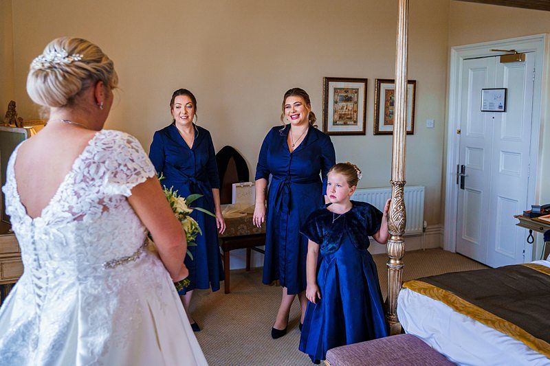 A bride in a white wedding dress and updo hairstyle stands in a room with three bridesmaids and a young girl, all dressed in dark blue dresses. They are smiling and appear to be enjoying a lighthearted moment together. The room has a four-poster bed and framed artwork. Image by Documentary Wedding Photographer Lewis Bishop Photography.