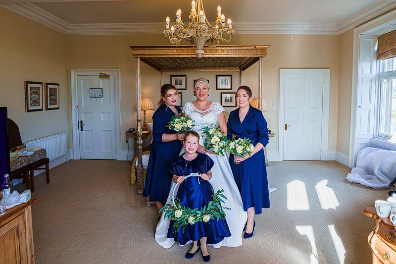 A bride stands with three bridesmaids in a well-lit room. The bridesmaids and a younger girl in front wear dark blue dresses and hold green and white floral bouquets. A chandelier hangs from the ceiling, and the room features elegant furniture and decor. Image by Documentary Wedding Photographer Lewis Bishop Photography.