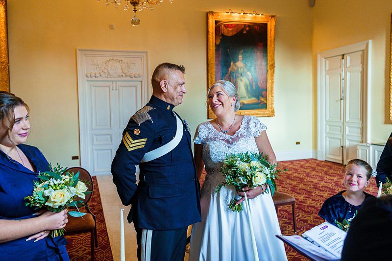 A bride and groom stand smiling at each other during their wedding ceremony in an elegant room with paintings on the walls. The groom is in a formal military uniform, and the bride is holding a bouquet of flowers. A smiling child and two women are also present. Image by Documentary Wedding Photographer Lewis Bishop Photography.