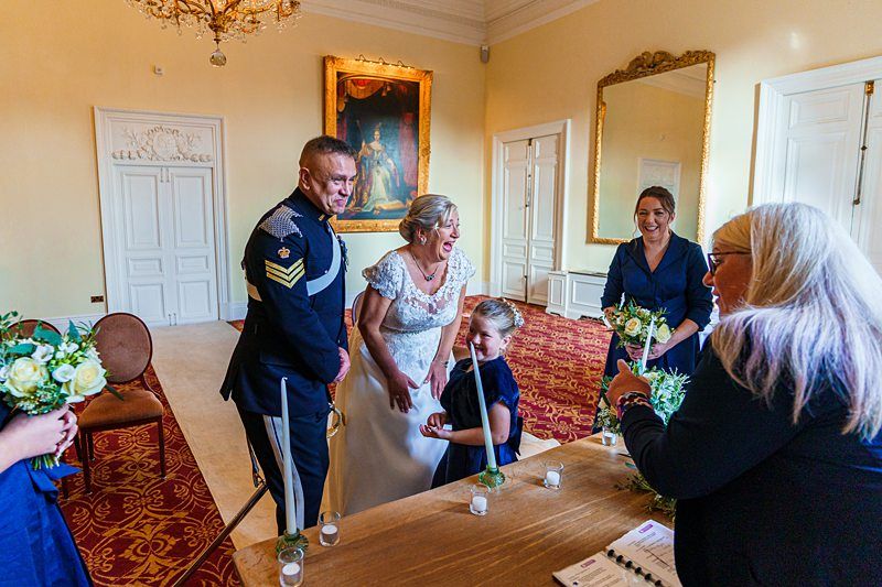 A bride in a white gown and a groom in a military uniform stand smiling with their daughter at a table in an elegant room with chandeliers and mirrors. Two women, one holding flowers, also smile nearby. Image by Documentary Wedding Photographer Lewis Bishop Photography.