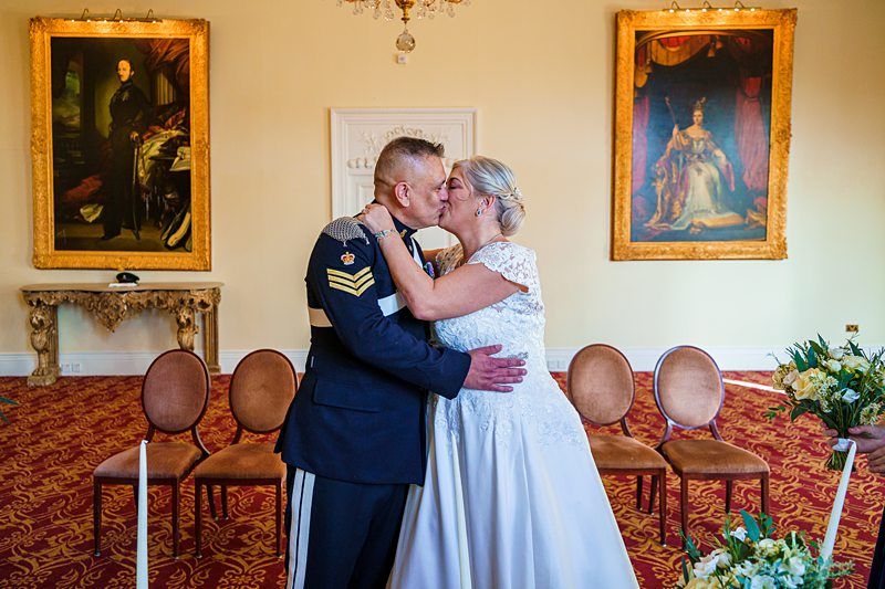 A couple dressed in formal attire share a kiss in a room adorned with two large framed portraits on the walls. The man is in a military uniform and the woman is in a white dress. They are embracing in front of several empty chairs and floral decorations. Image by Documentary Wedding Photographer Lewis Bishop Photography.