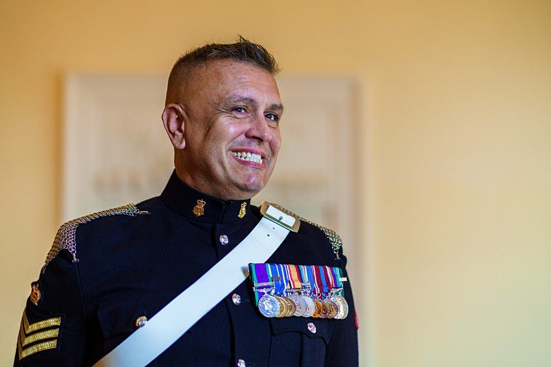 A person dressed in a formal military uniform stands smiling. They are wearing several medals on their chest and have short-cropped hair. The background is blurred, placing focus on the individual's bright expression and the detailed uniform. Image by Documentary Wedding Photographer Lewis Bishop Photography.