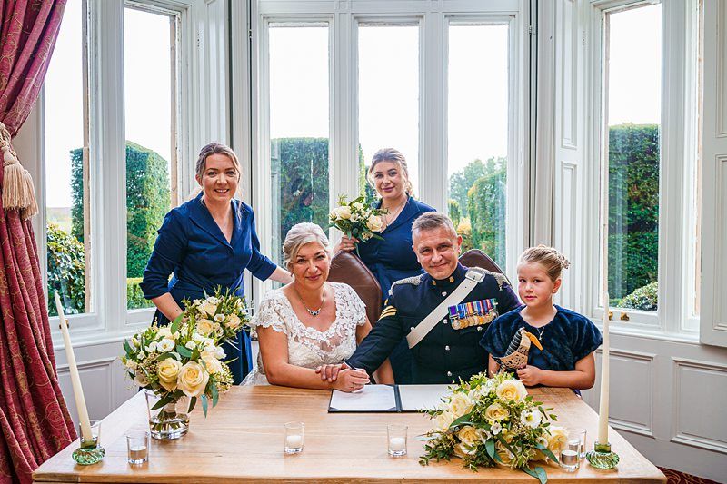 A bride in a white dress and groom in a military uniform sit at a table adorned with flowers, flanked by two adult women in blue dresses and a young girl in a navy dress. They are posed in front of large windows with greenery visible outside. Image by Documentary Wedding Photographer Lewis Bishop Photography.
