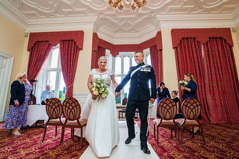 A bride and groom walk down the aisle in an elegant room with red curtains and an ornate ceiling. The bride wears a white gown and holds a bouquet, while the groom is dressed in a military uniform. Guests in the background look on and take photos. Image by Documentary Wedding Photographer Lewis Bishop Photography.