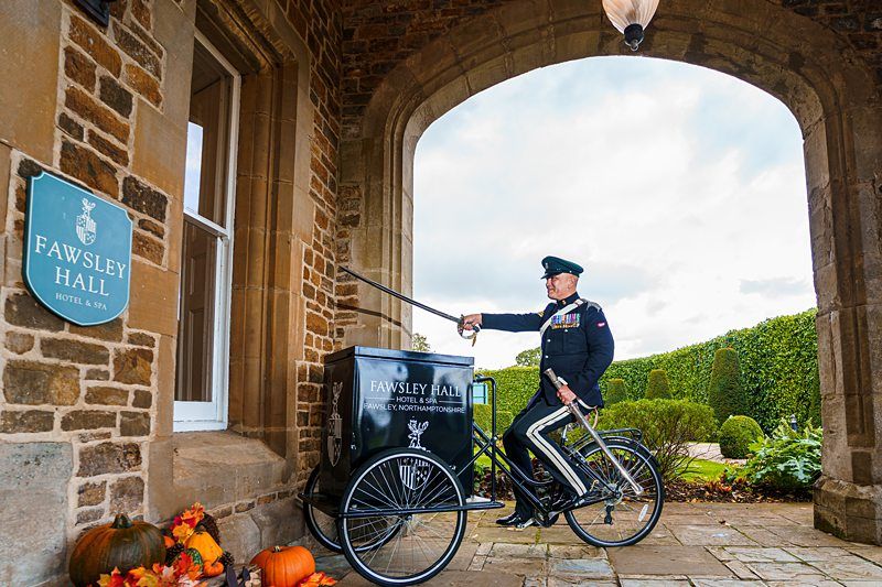 A uniformed man, standing under a stone archway at the entrance of Fawsley Hall Hotel & Spa, raises a sword while standing beside a vintage tricycle cart. Autumnal decorations, including pumpkins, are arranged on the ground nearby. Image by Documentary Wedding Photographer Lewis Bishop Photography.