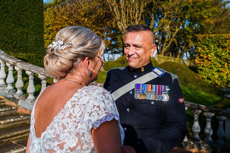 A man in a formal military uniform, adorned with medals, smiles at a woman in a white lace dress. They are standing outdoors near stone steps and balustrades, with trees displaying autumn foliage in the background. Image by Documentary Wedding Photographer Lewis Bishop Photography.