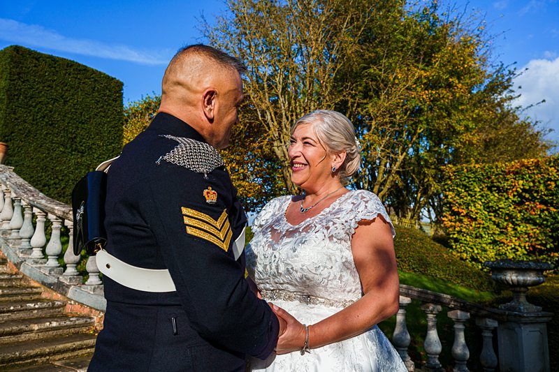 A happy couple, dressed in formal attire, stands outdoors on a sunny day. The man is in a dark military uniform with ornate details, while the woman wears a white lace wedding dress. They are holding hands and smiling at each other, surrounded by lush greenery and a stone staircase. Image by Documentary Wedding Photographer Lewis Bishop Photography.