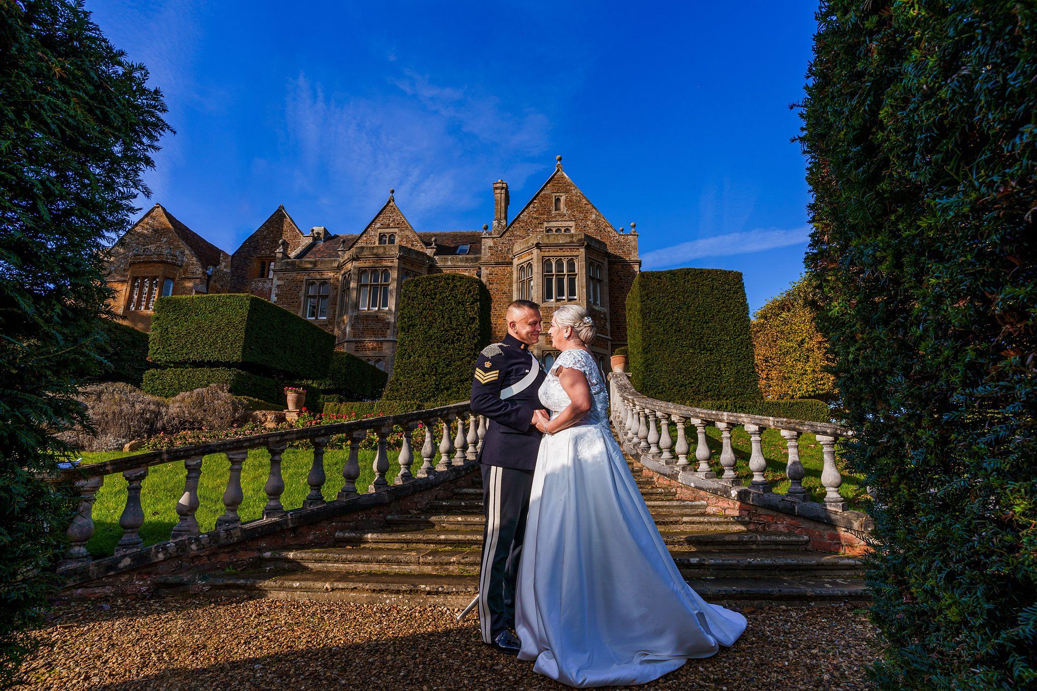 A bride in a white dress and a groom in a military uniform stand close together on a stone staircase outside a large, historic building with manicured greenery and a bright blue sky above. The couple embraces, creating a romantic scene in front of the grand estate. Image by Documentary Wedding Photographer Lewis Bishop Photography.
