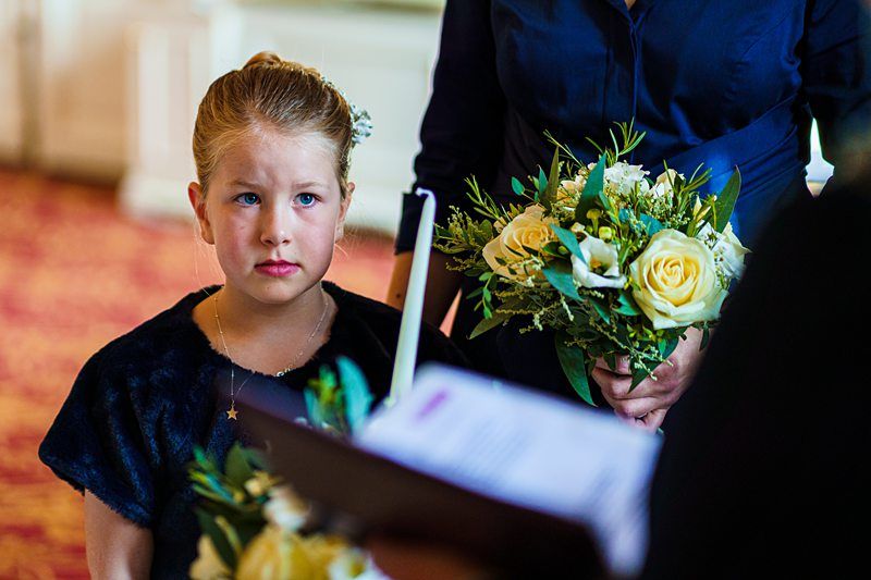 A young girl with blond hair tied back, wearing a dark dress, holds a floral bouquet and looks intently forward. Beside her, an adult in a navy outfit holds a similar bouquet. They are indoors with a red patterned carpet visible in the background. Image by Documentary Wedding Photographer Lewis Bishop Photography.