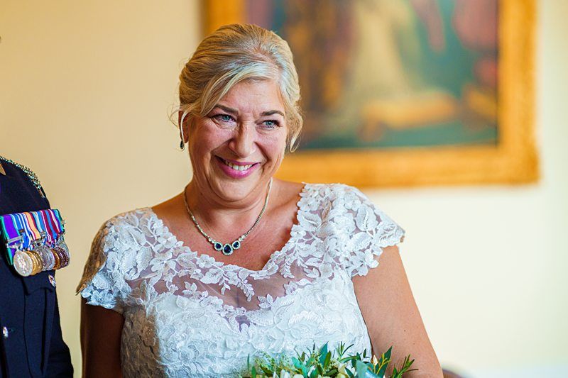 A smiling woman with blonde hair, wearing a white lace dress and a necklace with blue stones, holds a bouquet of greenery and white flowers. She stands beside someone uniformed with military medals. A framed painting is blurred in the background. Image by Documentary Wedding Photographer Lewis Bishop Photography.