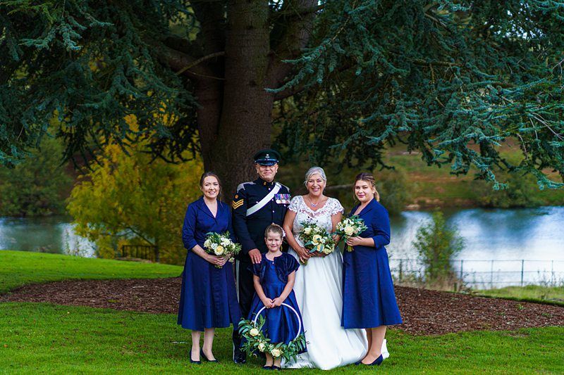 A group of five people is posing outdoors in front of a scenic backdrop. The woman in a white wedding dress is standing next to a man in a formal military uniform. They are surrounded by three women in blue dresses. Trees and a lake are visible in the background. Image by Documentary Wedding Photographer Lewis Bishop Photography.