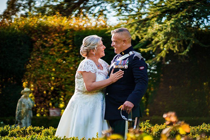 A smiling older couple stands in a lush garden, with the woman in a white wedding dress and the man in a formal military uniform adorned with medals. They are looking at each other affectionately, with sunlight filtering through the trees behind them. Image by Documentary Wedding Photographer Lewis Bishop Photography.
