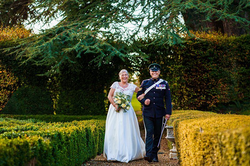 A bride in a white dress and a groom in a formal military uniform walk arm in arm down a path, smiling joyfully. They are surrounded by lush greenery and neatly trimmed hedges with sunlight filtering through the trees in the background. Image by Documentary Wedding Photographer Lewis Bishop Photography.