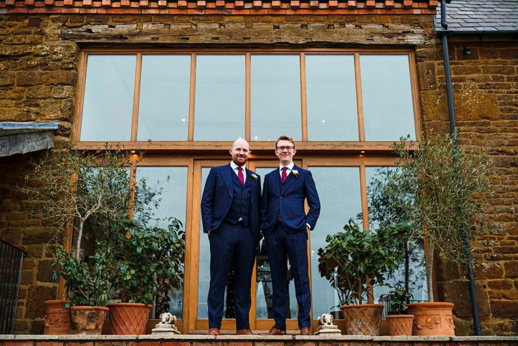 Two men in dark suits, wearing red ties and standing side by side, smile at the camera in front of a large modern window. The building has a rustic brick exterior. Potted plants and small statues are placed on the steps surrounding them. Image by Documentary Wedding Photographer Lewis Bishop Photography.