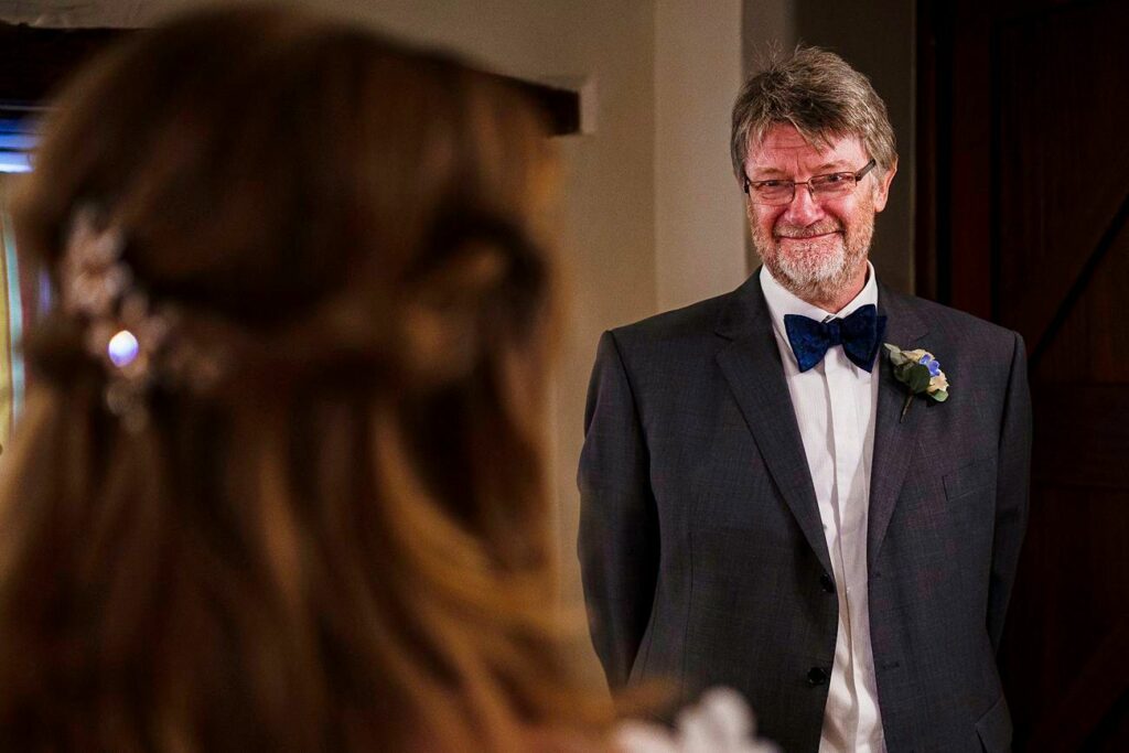 A man in a suit and bow tie, wearing glasses and smiling warmly, faces a woman with long hair, visible from behind. The setting appears to be indoors, and the man seems happy and proud as he looks at the woman. Image by Documentary Wedding Photographer Lewis Bishop Photography.